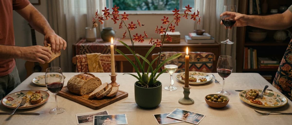 Mesa posta à noite: Wilsonara vermelha em vaso musgo no centro, velas acesas, taças de vinho, pão cortado e as mãos de duas pessoas.
